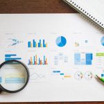 Overhead view of charts and graphs with magnifying glass and stationery on wooden table.